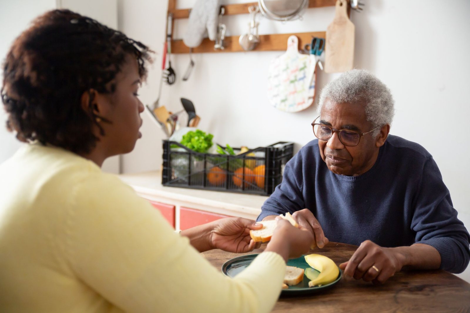 A Black caregiver setting a home-cooked plate of food on the kitchen table in front of an elderly Black woman in her own